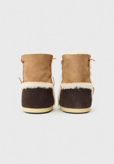 Botas de tobillo de ante marrón y beige con forro de piel sintética color crema, que presentan un detalle de cordones en el lado y una sección inferior de color marrón oscuro.