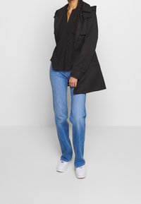 Černý trench kabát přes černé tričko, spárovaný se světle modrými džíny a bílými teniskami. Kabát má zapínání na knoflíky a pečlivě zpracovaný design.