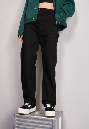 Personne portant un pantalon large noir, un crop top noir, une veste verte et des baskets à plateforme noires debout sur une boîte en métal argenté.