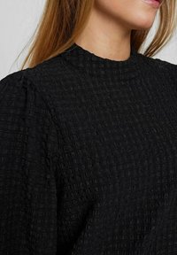 Zwarte blouse met een gestructureerd oppervlak, een opvallend vierkant patroon, hoge halslijn en puffmouwen. Gemaakt van een lichtgewicht stof.