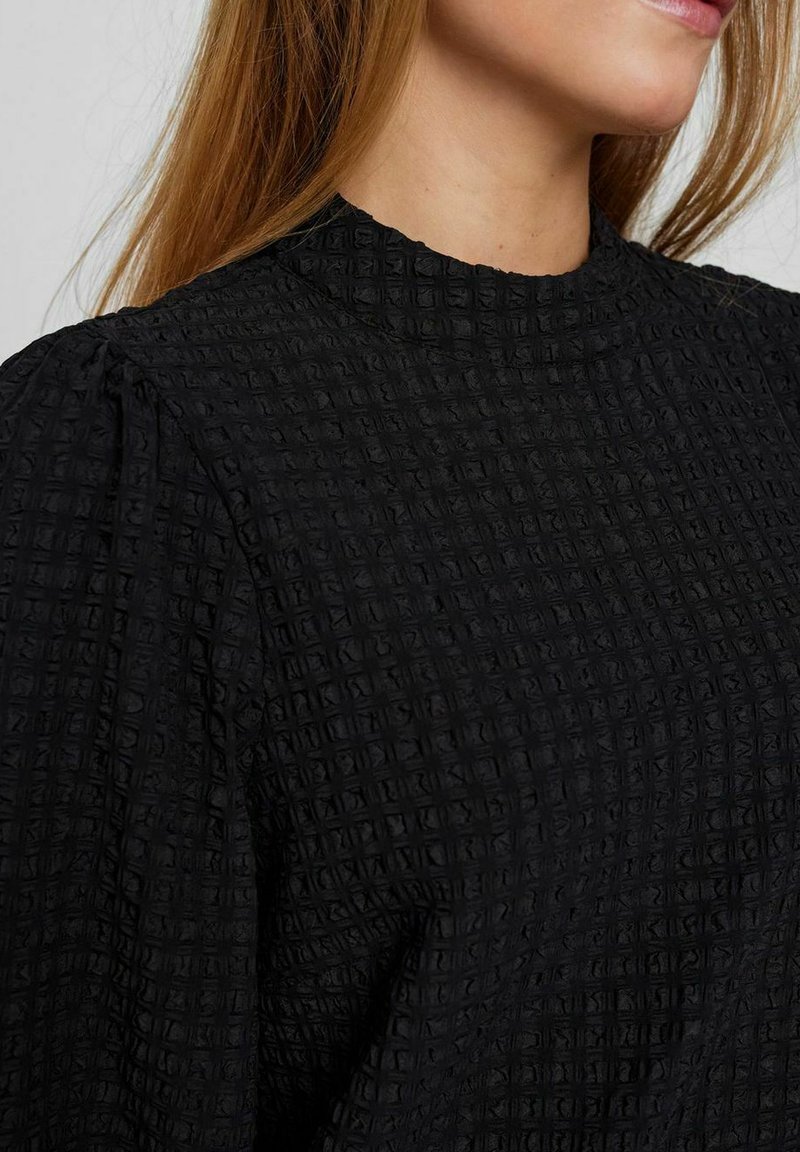 Czarna bluzka o teksturze z wyraźnym kwadratowym wzorem, wysokim dekoltem i bufiastymi rękawami. Wykonana z lekkiego materiału.