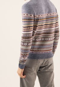 Knit sweater featuring multicolored geometric patterns, round neckline, and ribbed cuffs. Worn with gray pants, displaying a textured surface.
