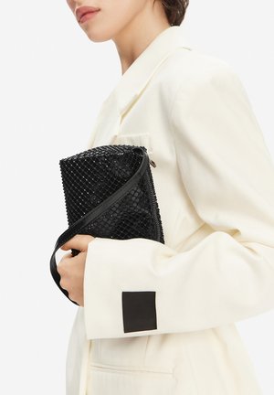 Žena v bílém saku drží u hrudi černou texturovanou obdélníkovou kabelku s dlouhým popruhem.