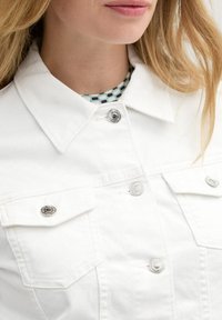 Veste en denim blanc avec des boutons-pression argentés et deux poches poitrine, dotée d'un col pointu et d'un fini en tissu texturé.