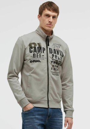 Young man wearing a light gray zip-up jacket with black printed text and blue jeans, standing with one hand in his pocket.