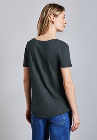 Tmavě zelené tričko s krátkým rukávem, kulatým výstřihem a volným střihem, vyrobené z měkké a lehké látky. Modré džíny dotvářejí tento outfit.