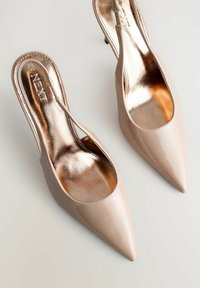 Tacones destalonados de charol nude con punta puntiaguda, forro interior metálico y tacón delgado. Presentan un diseño con recortes en los laterales.