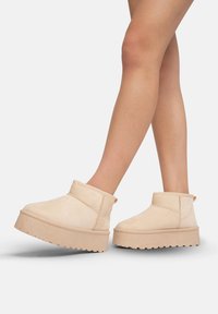Botines de plataforma beige con una suave textura de ante, puntera redondeada y detalles de costura. Cuenta con una lengüeta en la parte trasera para ponerlos y quitarlos fácilmente.