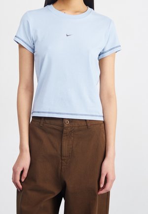 Person iført en lyseblå T-shirt med korte ærmer med Nike-logo og højtaljede løstsiddende brune bukser mod en ensfarvet baggrund.