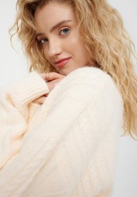 Kabelstrickpullover in sanftem Creme, mit gerippten Bündchen, strukturiertem Muster und entspannter Passform. Der Stoff wirkt gemütlich und warm.