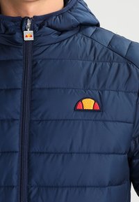 Námořnická puffer bunda s horizontálním prošíváním, s předním zipem a logem v kontrastních barvách červené a žluté.
