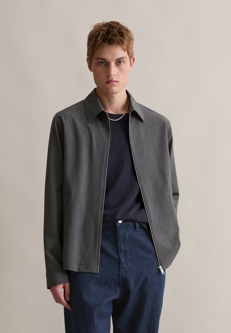 Szara kurtka na zamek z teksturowanym wzorem, szerokim kołnierzem i długimi rękawami, noszona na ciemnej koszulce, w połączeniu z niebieskimi spodniami.