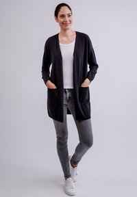 Cardigan lungo grigio scuro realizzato in un morbido materiale lavorato a maglia, con un design ampio, apertura frontale e due tasche anteriori. Indossato con jeans chiari.