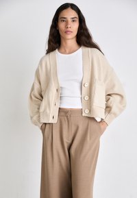 Beige gebreide cardigan met geribde manchetten, vier grote knopen en een zak, gecombineerd met losse bruine broeken en een witte top.