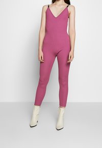 Kvinna i en åtsittande ärmlös mauve jumpsuit med V-ringning och vita ankelstövlar, stående mot en enkel ljus bakgrund.