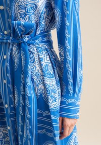 Abito camicia blu con stampa paisley bianca, con vita legata, chiusura anteriore a bottoni e maniche lunghe con dettagli a bottone. Tessuto morbido.