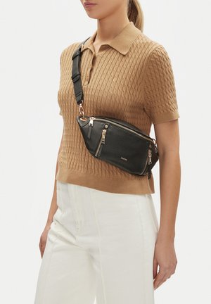 Mujer con polo de punto de manga corta color beige y pantalones blancos, con un bolso cruzado de cuero negro con cremalleras y herrajes dorados.