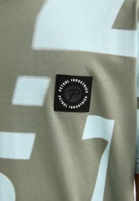 Zelené bavlněné tričko s světle modrými abstraktními tvary a černou štítkem s bílým textem a logem. Hladká textura, krátké rukávy.