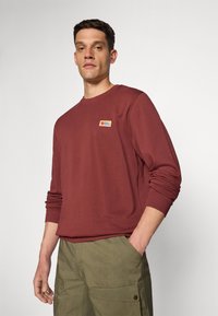 Burgunderfarget genser med rund hals og et lite flerfarget logo-patch på brystet. Har ribbede mansjetter og en avslappet passform.
