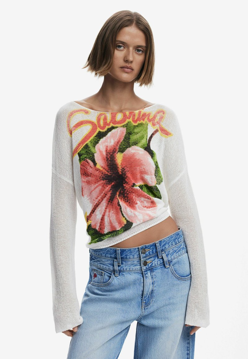 Maglione bianco lavorato a maglia con un grande design floreale di ibisco rosa e il testo "Sabrina" sul davanti. Abbinato a jeans blue chiaro a vita alta.