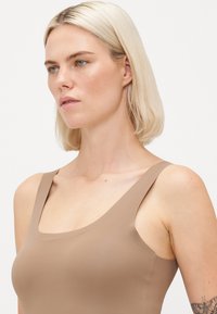 Blonde woman with short hair wearing a beige sleeveless top, looking thoughtfully to the left against a plain white background.