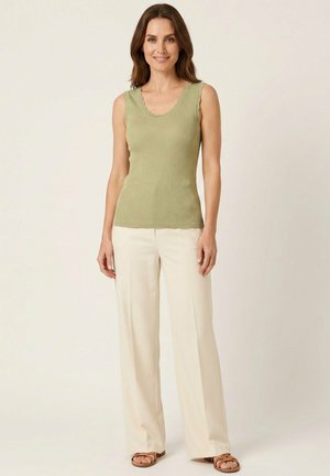 Femme portant un haut en tricot sans manches vert clair, un pantalon blanc à jambes larges et des sandales marron, debout devant un fond beige uni.