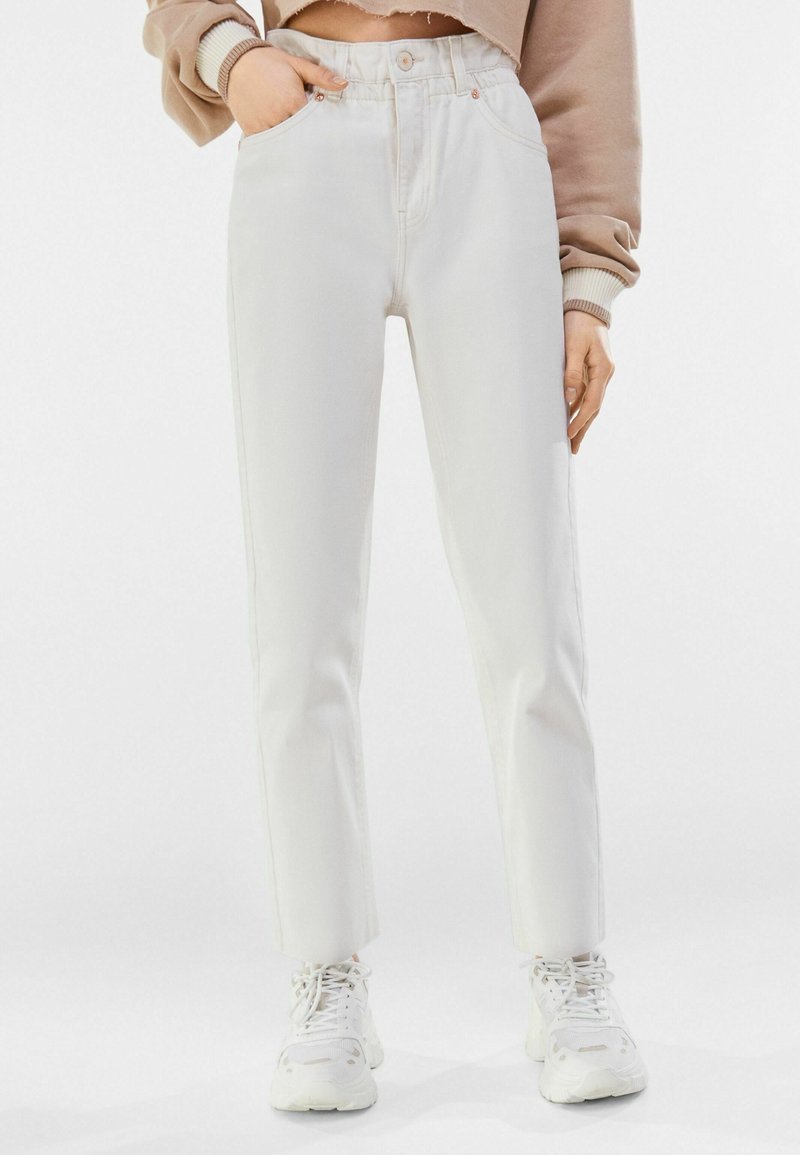 Pantalones de mezclilla blancos con cintura alta, piernas ajustadas, bolsillos frontales y cierre de botón, combinados con zapatillas blancas. La textura parece suave.