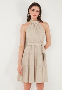 Vaalean beige halterneck-mekko, jossa on muotoon ommeltu vyötärö, kerroksellinen helma ja kankainen solmu vyötäröllä. Sileä rakenne ja hienovarainen laskostus.