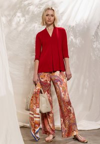 Blouse rouge à manches longues avec col en V et design en couches, associée à un pantalon à jambes larges à motif cachemire orange, des sandales dorées et un sac à main beige.