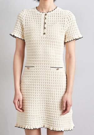 Femme portant une robe courte en maille crème avec des boutons sur le devant et des bords festonnés bordés de noir aux manches et à l'ourlet.