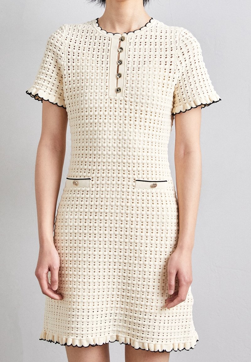 Femme portant une robe courte en maille crème avec des boutons sur le devant et des bords festonnés bordés de noir aux manches et à l'ourlet.