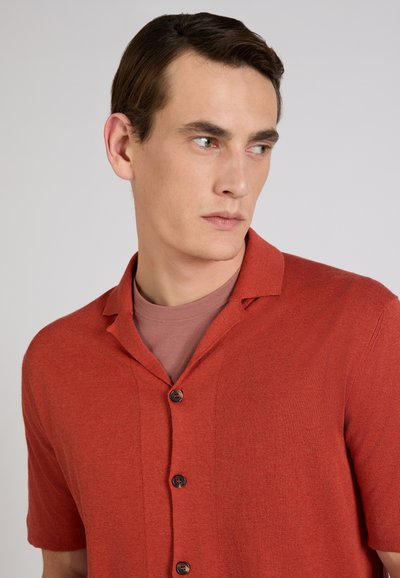 Jeune homme aux cheveux courts et foncés portant une chemise à manches courtes boutonnée couleur rouille par-dessus un t-shirt col rond mauve pâle, regardant sur le côté.