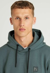 Groene hoodie met een textuurafwerking, voorzien van een voorste kangoeroezak, zwarte trekkoorden en een klein logo-embleem op de borst.