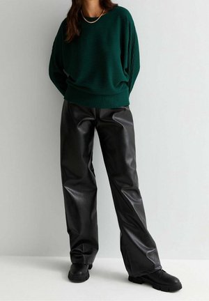 Žena v zeleném žebrovaném svetru, černých širokých kalhotách z umělé kůže, černých robustních botách a zlatém náhrdelníku na jednoduchém pozadí.