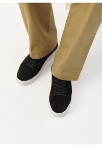 Personne portant des baskets noires avec des semelles blanches et un pantalon beige, debout sur une surface blanche.