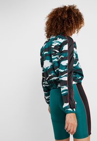 Kamouflagemönstrad jacka i teal, svart och vitt med elastisk midja; kombinerad med teal-korta byxor som har en svart och röd sidrand.