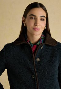 Giacca in pile blu scuro con colletto in velluto a coste marrone, dotata di un laccio e due grandi bottoni. Sopra una camicia a quadri.