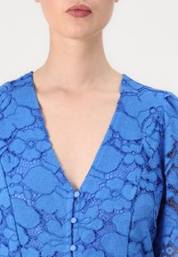 Blue lace dress with a v-neckline, decorative buttons, and textured floral patterns. Features a blend of opaque and sheer fabrics.