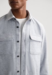 Jasnoszara flanelowa koszula z czarnymi guzikami, dwoma kieszeniami na piersi i kołnierzykiem. Miękka tekstura i luźny krój.