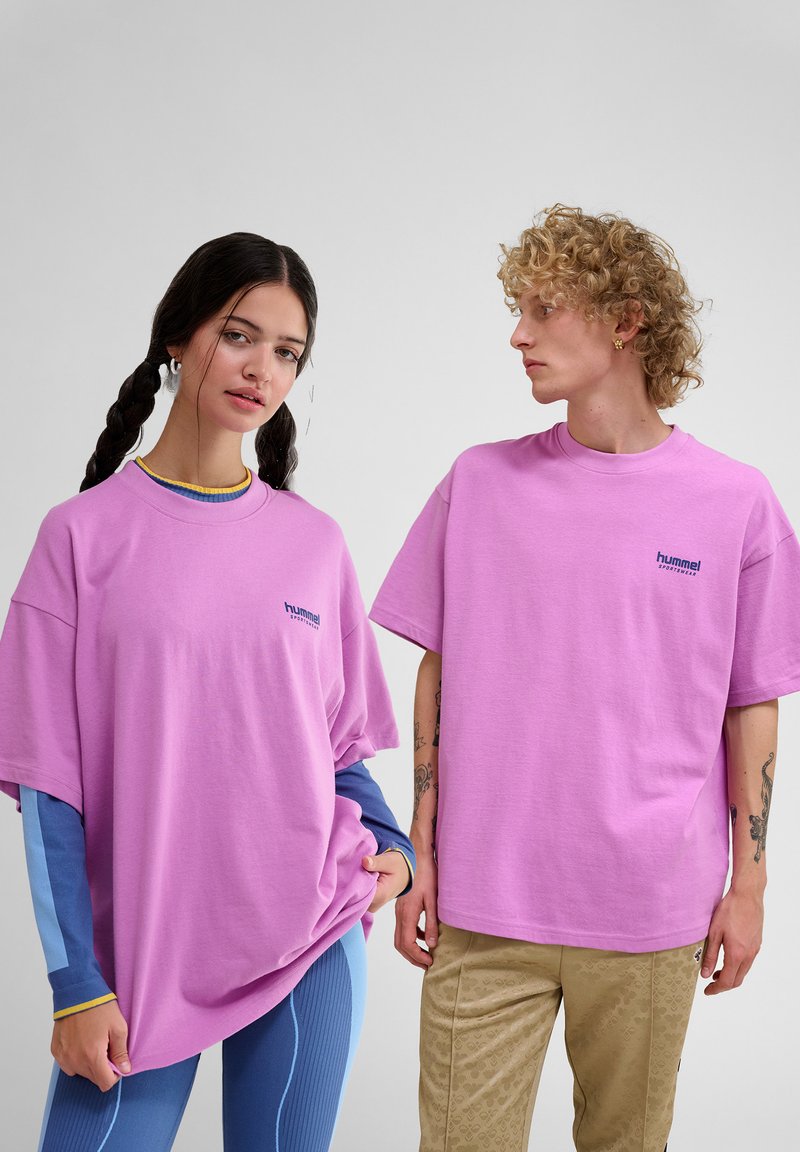 To personer iført oversized lyserøde t-shirts med et lille logo. T-shirtsne har en blød tekstur og en afslappet pasform, stylet med kontrasterende lag.