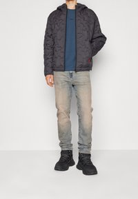 Gepolsterte schwarze Kapuzenjacke mit Reißverschluss, getragen über einem blauen T-Shirt, kombiniert mit hellen Jeans und schwarzen Turnschuhen.