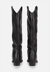 Botas altas de cuero negro con textura perforada, punteras afiladas y un diseño ligeramente caído. Tacón bajo apilado con acabado suave.