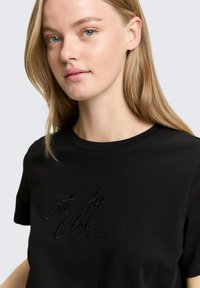 Schwarzes Baumwoll-T-Shirt mit Rundhalsausschnitt und kurzen Ärmeln, das auf der Vorderseite ein erhabenes Design "5th Ave Club" aufweist.
