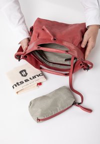 Rode leren tas met een stoffen voering, voorzien van een rits en meerdere klinknagels. Inclusief een beige katoenen pouch met een logo en een klein groen ritsvakje.