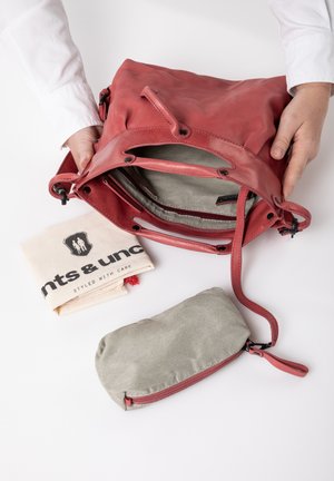 Rode leren tas met een stoffen voering, voorzien van een rits en meerdere klinknagels. Inclusief een beige katoenen pouch met een logo en een klein groen ritsvakje.