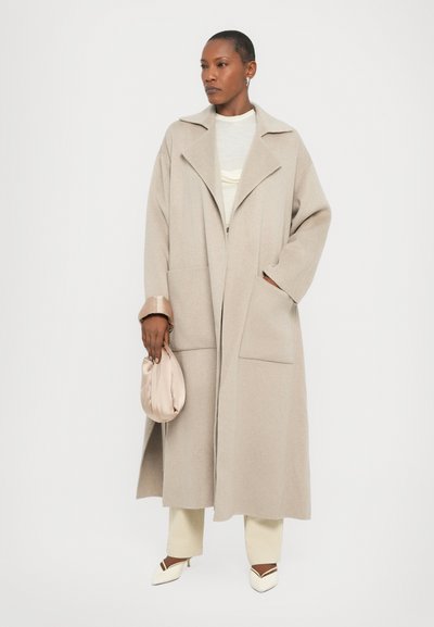 Cappotto di lana beige con un ampio colletto, due tasche frontali e maniche larghe, abbinato a pantaloni crema e scarpe bianche a punta.