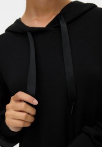 Černá mikina s kapucí a šňůrkou, ribbené manžety. Měkká textilie, uvolněný střih a minimalistický design. Detailní záběr na ruce držící šňůrky.