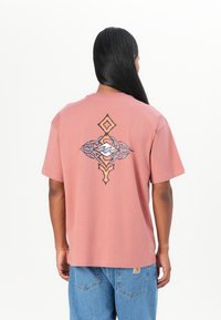 Pink cotton t-shirt with a loose fit, featuring a detailed graphic design in black, orange, and white on the back. Short sleeves, crew neck.