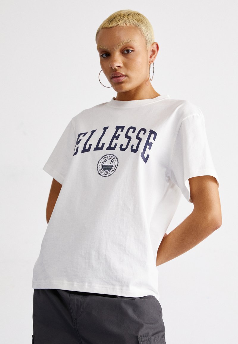 Camiseta de algodón blanca con el texto en negrita "ELLESSE" en azul marino y logo circular en el centro. Corte relajado, mangas cortas, textura suave.