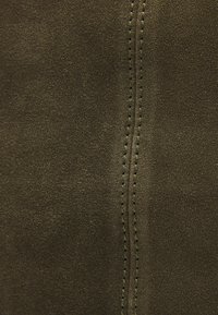 Tissu en suède vert olive avec une texture lisse, doté d'une double couture dans une teinte légèrement plus foncée. Les points de couture sont serrés et réguliers.